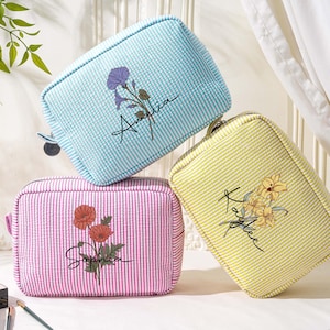 May include: Three rectangular cosmetic bags in pastel colors: light blue, pink, and yellow. Each bag features a floral design and a name in script. The bags have a seersucker texture and a zipper closure, ideal for storing makeup and toiletries.