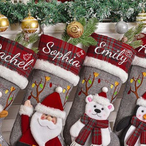 May include: Four personalized Christmas stockings with names Rachel, Sophia, Emily, and Amelia embroidered in white. Each stocking has a red and gray plaid cuff and is decorated with festive characters like a reindeer, Santa, a polar bear, and a penguin.