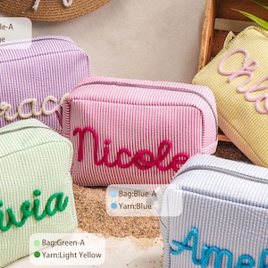 May include: Five rectangular zippered pouches in pastel striped fabric, each featuring a name embroidered in raised yarn. The bags are purple with beige text, pink with dark pink text, yellow with pink text, green with green text, and blue with blue text.