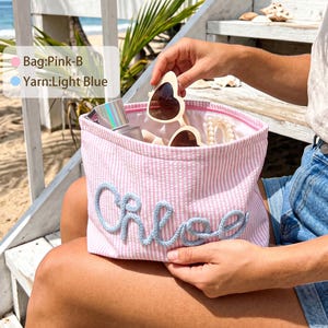 May include: A pink and white striped zippered pouch featuring the name Chloe in light blue yarn lettering. The pouch contains heart-shaped sunglasses, a small bottle, and a hair accessory. Text on the image reads Bag:Pink-B and Yarn:Light Blue.