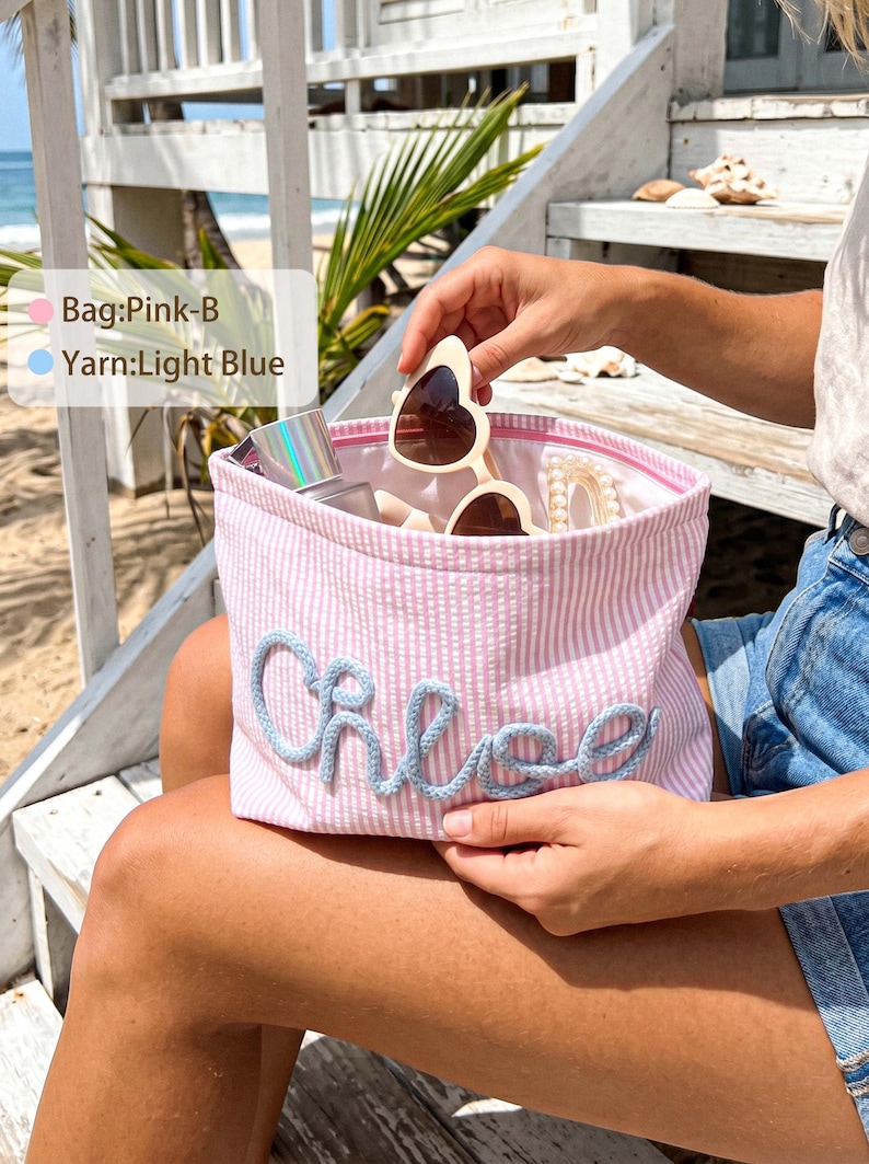 May include: A pink and white striped zippered pouch featuring the name Chloe in light blue yarn lettering. The pouch contains heart-shaped sunglasses, a small bottle, and a hair accessory. Text on the image reads Bag:Pink-B and Yarn:Light Blue.