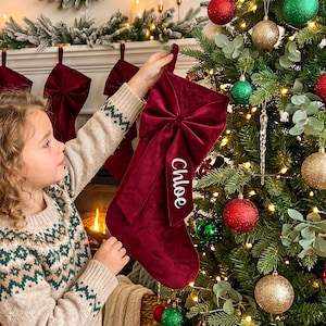 May include: A deep red velvet Christmas stocking featuring a large, matching velvet bow on the front. The stocking has a long, dangling fabric tag with the name Chloe printed in white script lettering. The material has a soft, plush texture.