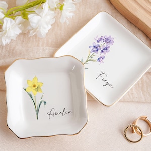 Op de afbeelding: Twee witte keramische ringbakjes met gouden rand. E&eacute;n bakje heeft een gele narcis en de naam "Amelia". Het andere bakje heeft paarse bloemen en de naam "Freya". Gouden ringen liggen naast de bakjes. Witte bloemen op de achtergrond.