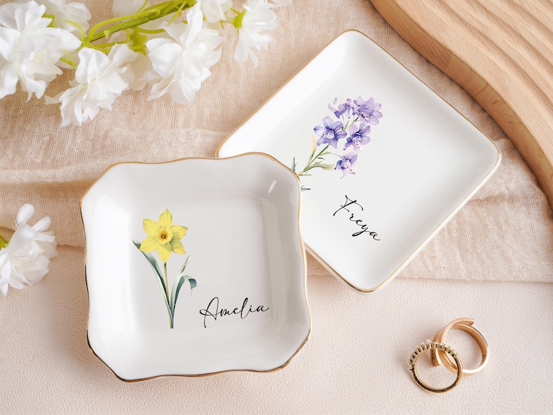 Op de afbeelding: Twee witte keramische ringbakjes met gouden rand. E&eacute;n bakje heeft een gele narcis en de naam "Amelia". Het andere bakje heeft paarse bloemen en de naam "Freya". Gouden ringen liggen naast de bakjes. Witte bloemen op de achtergrond.