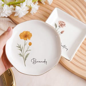 Op de afbeelding: Twee witte keramische schalen met gouden randen. E&eacute;n is rond met een oranje bloem en de naam "Brandy". De andere is vierkant met een roos en het woord "Destiny". Beide hebben bloemmotieven.