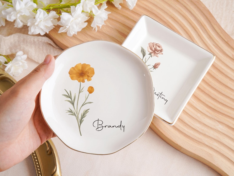 Op de afbeelding: Twee witte keramische schalen met gouden randen. E&eacute;n is rond met een oranje bloem en de naam "Brandy". De andere is vierkant met een roos en het woord "Destiny". Beide hebben bloemmotieven.