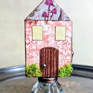 May include: A whimsical miniature house with a pink floral pattern, a wooden door, and two yellow-windowed openings. The roof features mushroom illustrations. The house is mounted on a clear, faceted glass base, with green moss accents.