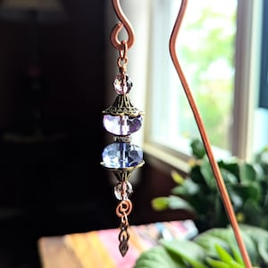 May include: A handcrafted copper bookmark with a decorative beaded charm. The charm features faceted purple and clear beads, accented with antique brass-colored metal elements. The bookmark is suspended in front of a window with greenery.