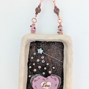 May include: A handmade wall hanging featuring a rectangular frame with a dark, glittery background. A lavender heart with the word "Love" is centered, with a silver star wand and sparkling accents. The piece is adorned with beaded copper wire.
