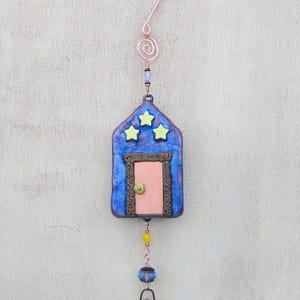 May include: A whimsical, handmade blue house ornament with three yellow stars, a pink door, and a copper wire hanger. The ornament is adorned with beads and a small basket holding a crystal. The overall design is artistic and decorative.