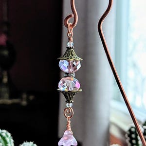 May include: A handcrafted copper bookmark with a decorative design. The bookmark features two faceted, iridescent crystal beads and a small, light pink flower charm. The top of the bookmark has a looped design.