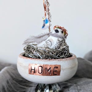 May include: A decorative bird figurine in a nest, resting in a white bowl with a copper "HOME" plaque. The bird has a scarf and a floral crown. A crescent moon charm hangs above. The bowl sits on a crystal base.