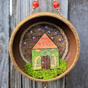 May include: A circular wooden frame containing a miniature house with a green patterned facade and a brown door. The house sits on green moss against a background of a vintage map with iridescent star accents. It hangs from copper wire with red beads.