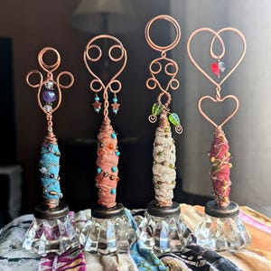 May include: Four decorative stands featuring clear glass bases and copper wire tops. Each stand has a fabric-wrapped center adorned with beads. The wire tops include various shapes like circles and hearts, accented with small charms and crystals.