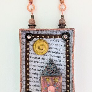 May include: A handcrafted rectangular pendant with a house design. The pendant features a small house with a pink facade and a blue roof, set against a backdrop of printed text. The frame is adorned with copper accents and decorative elements, and it hangs from a fabric-wrapped handle.