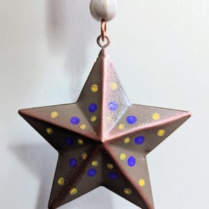 May include: A five-pointed star ornament with a metallic bronze finish, decorated with small hand-painted yellow and blue polka dots. The star hangs from a copper-colored jump ring attached to a white bead and a patterned fabric-covered top piece.