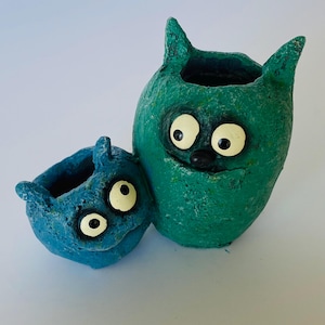 May include: Two whimsical, handmade ceramic planters. One is teal, the other is blue. Both have large, cartoonish eyes, pointed ears, and a textured surface. The planters are designed to resemble friendly monsters.