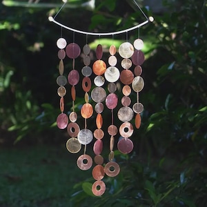 May include: A decorative wind chime with a variety of shapes and colors. The wind chime is made of circular and oval-shaped pieces in shades of copper, gold, silver, and purple. The pieces are strung together and suspended from a curved, white metal bar.