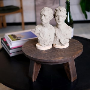 Könnte beinhalten: Zwei kleine, hellfarbene Tonbüsten von menschlichen Figuren stehen auf einem dunklen Beistelltisch aus Holz mit drei Beinen. Der Tisch steht auf einem größeren schwarzen Tisch. Ein Stapel Bücher und eine grüne Flasche befinden sich im Hintergrund.