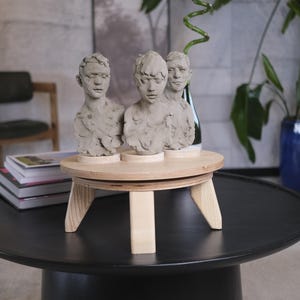 May include: Three clay busts of human figures sit on a small, round, wooden table with angled legs. The table is on a black, round coffee table. The busts are light gray and have detailed facial features. The background includes a chair, books, and plants.