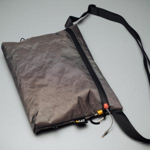 May include: A gray, rectangular shoulder bag with a black zipper and strap. The bag has a small carabiner clip and a tag that reads "CAT". The bag is made of a lightweight, textured material.