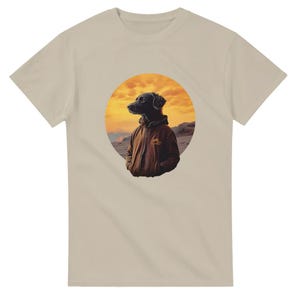 May include: A beige t-shirt featuring a circular graphic of a black dog wearing a brown jacket. The dog is standing on a beach with a sunset in the background.