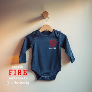 May include: A navy blue baby bodysuit with long sleeves, displaying a red firefighter emblem and the name "COOPER." The garment hangs on a wooden hanger. Text on the image reads "CUSTOM FIRE DEPARTMENT (On the back)."