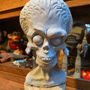 May include: A white painted bust of an alien with a large head and a detailed brain sculpted on the top. The alien has large eyes and a wide smile. The bust is sitting on a wooden base.