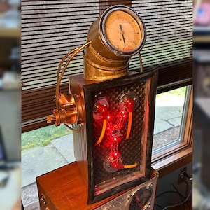 May include: A steampunk-style sculpture featuring a brass gauge, copper piping, and a rectangular frame with red lights. The sculpture is mounted on a wooden base, showcasing a unique industrial aesthetic.