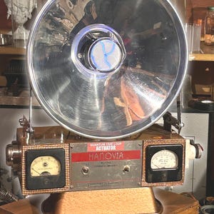 May include: A vintage Hanovia Alpine Travel Model device featuring a large, circular, polished chrome reflector bowl with a central blue glowing element. The base includes two analog gauges, a toggle switch, and a label reading Quantum Time Loop Actuator.