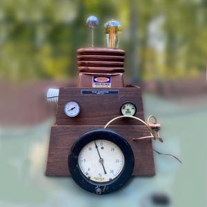 May include: A steampunk-style device with a copper top and wooden base. It features a light bulb, a small sphere, and gauges. The device has a large, round gauge with a black face and white numbers, and the text "DANGER HIGH VOLTAGE".