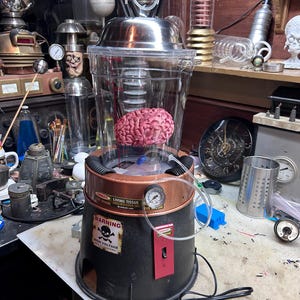 May include: A science fiction prop featuring a pink brain inside a clear plastic container with a metal lid. The black base has a copper-colored ring, a pressure gauge, a toggle switch, and warning labels that read DANGER LIVING TISSUE and WARNING HIGH VOLTAGE.