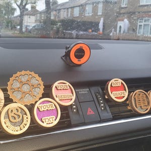 May include: A selection of wooden car air fresheners with various designs and text, including "Trinity", "Your Design", "Any Shape", and "Your Text". The air fresheners are shown on a car dashboard with a black phone holder.