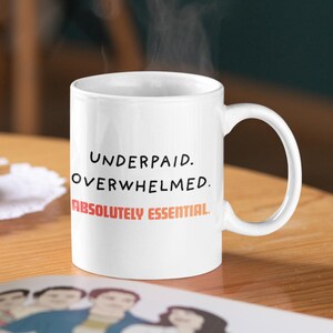 May include: White ceramic mug with the text "UNDERPAID. OVERWHELMED. ABSOLUTELY ESSENTIAL." in black and red lettering. The mug has a curved handle and is on a wooden surface. Steam rises from the mug.