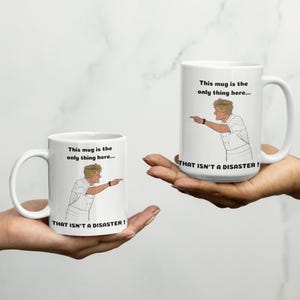 May include: Two white ceramic mugs featuring a cartoon illustration of a chef pointing and the text: This mug is the only thing here... THAT ISN'T A DISASTER! The design is printed on the front of both mugs, which have a standard handle.