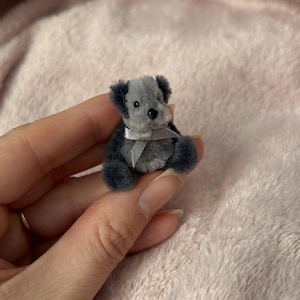 Puede incluir: Un pequeño oso de peluche gris con una cinta plateada alrededor del cuello. El oso está sentado en la mano de una persona.