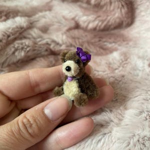 Pode incluir: Um pequeno urso de peluche castanho com um laço roxo na cabeça e um colar roxo. O urso está sentado numa mão.