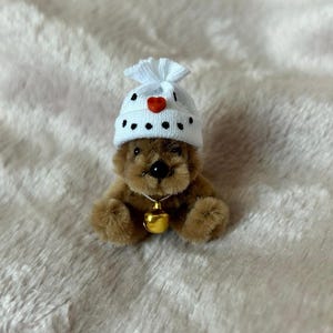 Pode incluir: Um pequeno urso de peluche castanho a usar um chapéu branco de malha com pontos pretos e um coração vermelho. O chapéu tem um pompom no topo. O urso usa um colar com um sino dourado.