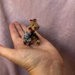 Può includere: Una miniatura di giraffa e koala marrone e grigia con dettagli di nastro verde. I piccoli peluche sono tenuti in una mano su uno sfondo rosa.