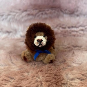 Puede incluir: Un pequeño juguete de peluche con forma de león, con una melena marrón y un cuerpo beige. El león tiene una cara blanca con una nariz y boca cosidas en negro. Una cinta azul está atada alrededor de su cuello. El juguete está sobre una superficie rosa y difusa.