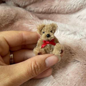 May include: A small, brown teddy bear with a red ribbon around its neck. The bear is sitting in the palm of a hand.