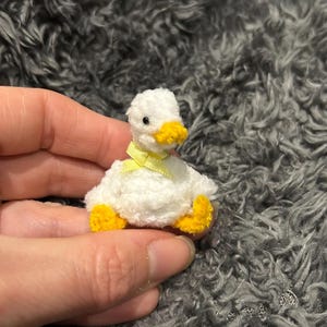 May include: A small white plush duck with yellow feet and a yellow bow around its neck. The duck is sitting on a gray fuzzy surface.