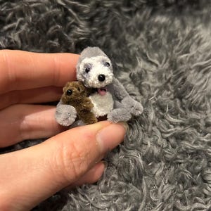 May include: A gray and white plush toy dog holding a brown plush toy bear. The dog has a pink nose and black eyes.