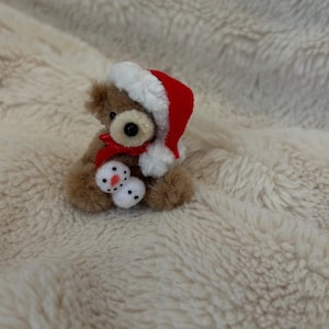 May include: A miniature brown teddy bear, adorned with a red and white Santa hat and scarf, clutching two tiny snowmen. Set against a soft, cream-coloured fabric backdrop.