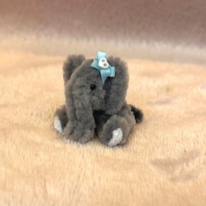 Peut inclure: Un petit éléphant en peluche gris avec un nœud bleu clair sur la tête. L'éléphant a des accents blancs sur les pieds et est assis sur une surface douce de couleur pêche. Le jouet est un article mignon fait à la main.