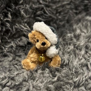 Pode incluir: Um pequeno urso de pelúcia marrom usando um chapéu branco e azul e um colar com um sino dourado. O urso está sentado em uma superfície cinza e difusa. A pelagem do urso é marrom claro.