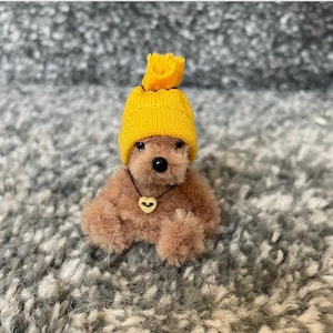 Puede incluir: Un pequeño oso de peluche marrón que lleva un gorro de punto amarillo y un collar con un colgante en forma de corazón.