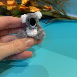 Könnte beinhalten: Ein Miniatur-Koala-Bär-Plüschtier in Grau mit weißen Akzenten an Ohren und Brust. Der Koala hat eine schwarze Nase und kleine, farbenfrohe herzförmige Verzierungen um den Hals. Das Plüschtier wird in einer Hand vor blauem Hintergrund gehalten.
