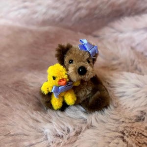 May include: A brown teddy bear with a blue bow is holding a yellow chick with a blue bow. The toys are on a light brown fluffy surface.