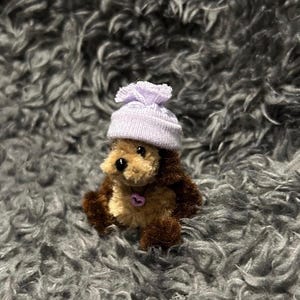 Pode incluir: Um cão de pelúcia em miniatura com pelo castanho e um chapéu lilás claro. O cão tem um pequeno pingente em forma de coração rosa. O brinquedo está sobre um fundo cinza e difuso.